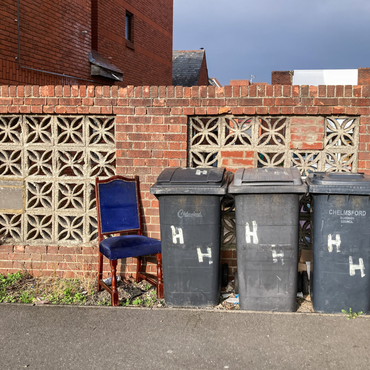 Photograph of 3 black wheelie bins with 'HH' painted on them in white, with an old-fashioned dark wood and blue velvet dining chair to the left. Behind is a brick wall with patterned square bricks, there's a pavement in front and an ominous looking sky above.