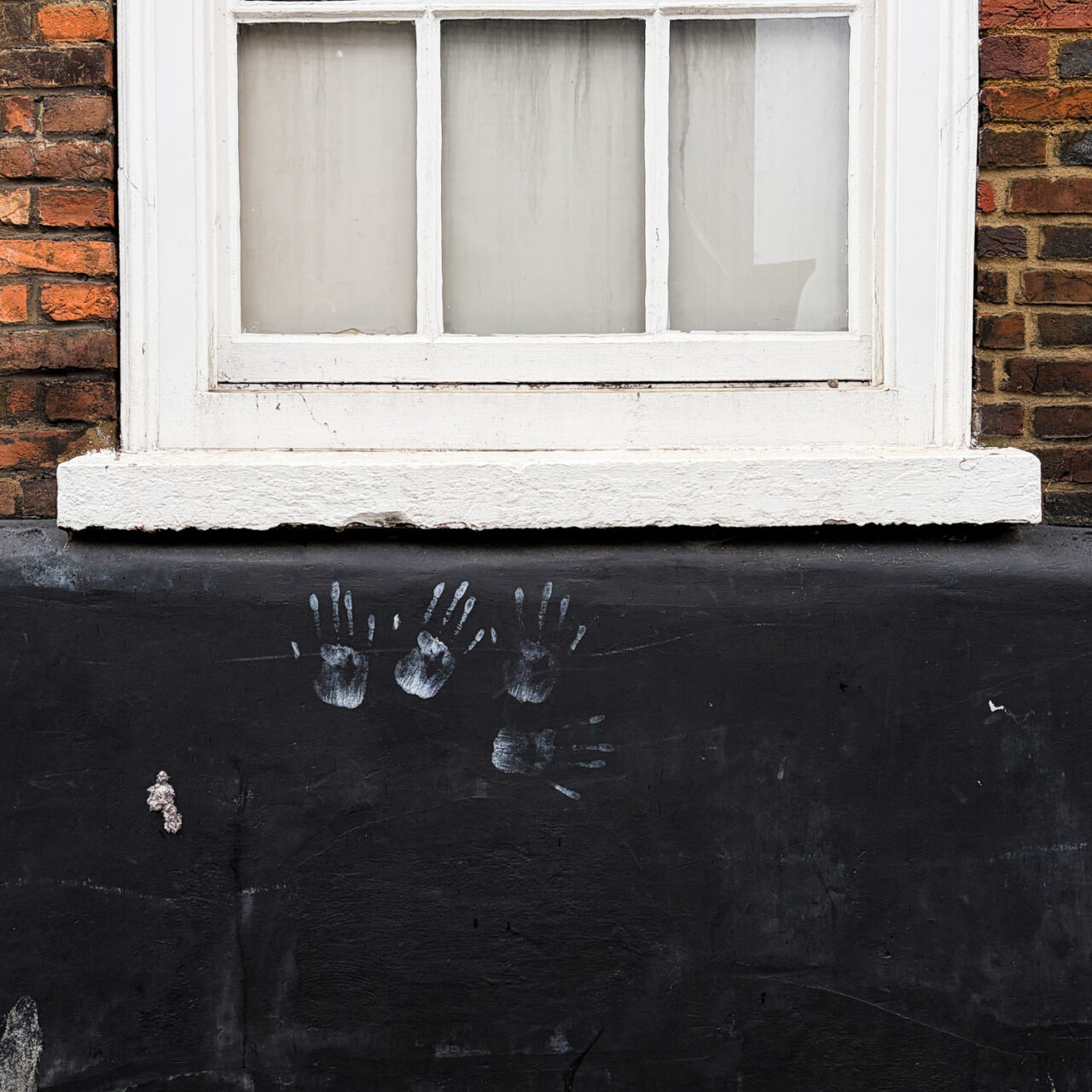 Photograph of a wall, which is painted black in the bottom half, with 4 white handprints clustered near the centre. Above is a brick wall with a white framed sash window above.